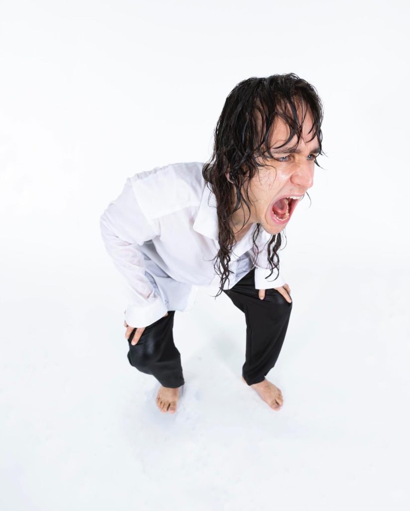 Yel est photographié depuis un point en hauteur sur un fond blanc. Iel semble crier. Ses cheveux sont longs, bruns et mouillés. Iel porte une chemise blanche et un pantalon noir et est pieds nus.